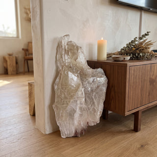 Selenite Window Slab Unique #16 - 30" displayed leaning against a wall on a light oak floor beside a lit candle, shown at true size in warm natural light.