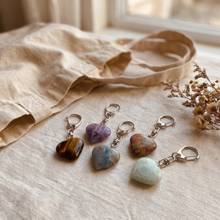 The five mixed stone heart charms laid on cream linen beside a canvas tote in soft natural light, shown at true scale.