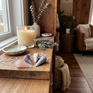 Mixed Stone Pendulums 10-Pack displayed on a wooden home altar shelf beside a lit candle and dried lavender, shown at true size in warm natural light.