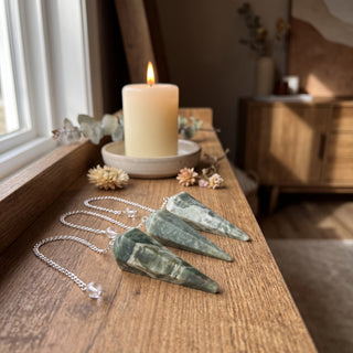 Vessonite Pendulum 6 Facet & Bead - 3 Pack on a wooden altar shelf beside a candle, three mottled green vessonite pendulums at true size in warm natural light.