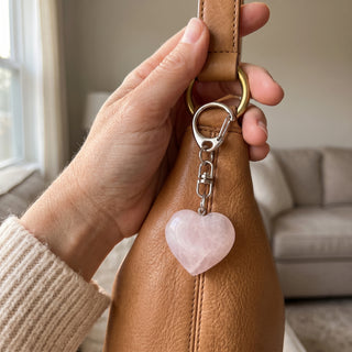 One of the five soft pink rose quartz heart charms from the pack, clipped to a tan leather handbag in natural light, shown at true scale.