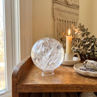 Clear Quartz Sphere Unique #19 - 6" displayed on a wooden home altar shelf beside a candle, shown at true size in warm natural light.