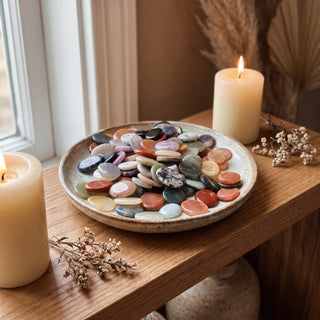 Mixed Stone Worry Stones 100-Pack displayed in a ceramic dish on a wooden home altar shelf beside a lit candle, shown at true size in warm natural light.