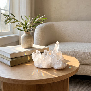 Clear quartz cluster glowing in afternoon sunlight on a white-oak side table beside linen books and an olive branch in a warm modern living room